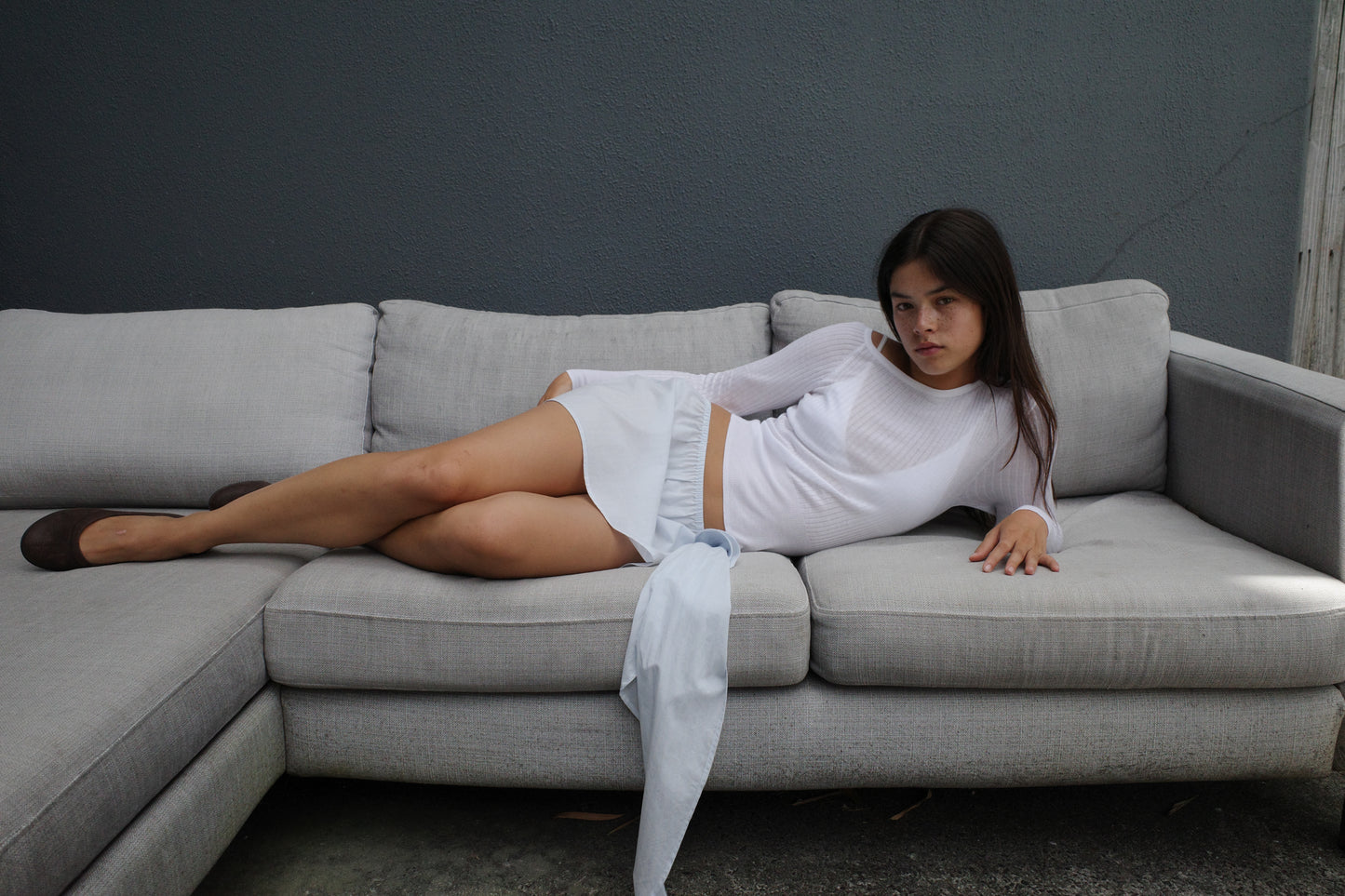 A young woman in a Deiji Studios Shoulder Knit Top - White lounges on a light gray sectional sofa against a dark gray wall, with one arm resting on the back and her legs stretched out on the cushions.