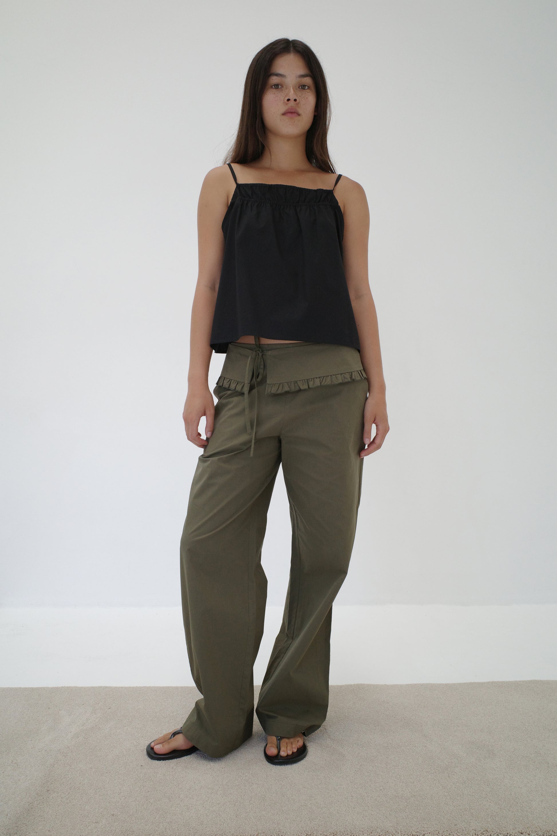 A woman with long dark hair and a neutral expression stands against a white wall wearing a loose black sleeveless top, black sandals, and relaxed fit Flounce Belt Pant - Khaki with ruffle detail and drawstring.