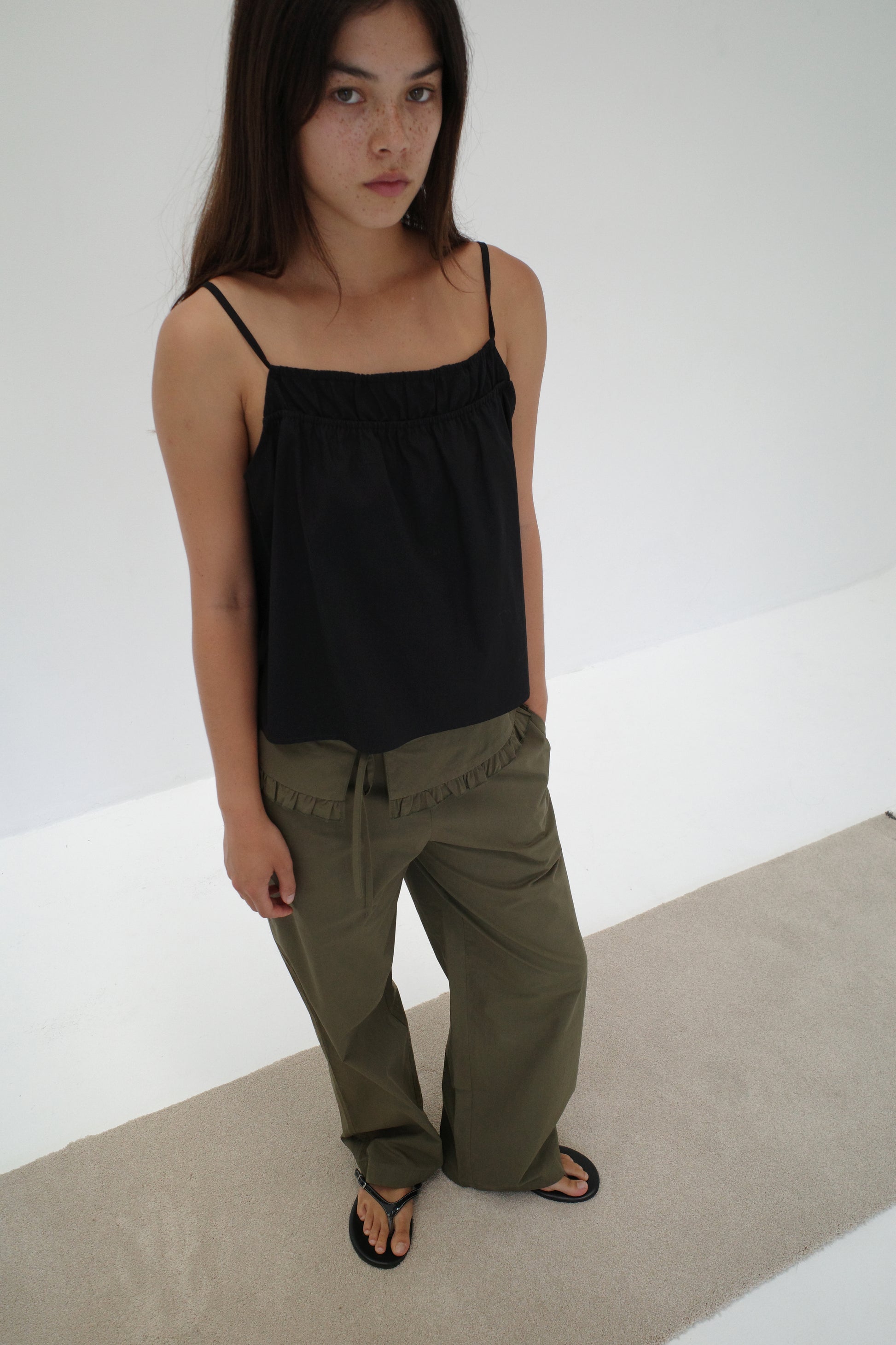 A woman with long brown hair stands on a light beige carpet, wearing the Flounce Belt Pant - Khaki, a black spaghetti strap top, and black sandals. She looks slightly up toward the camera against a plain white background.
