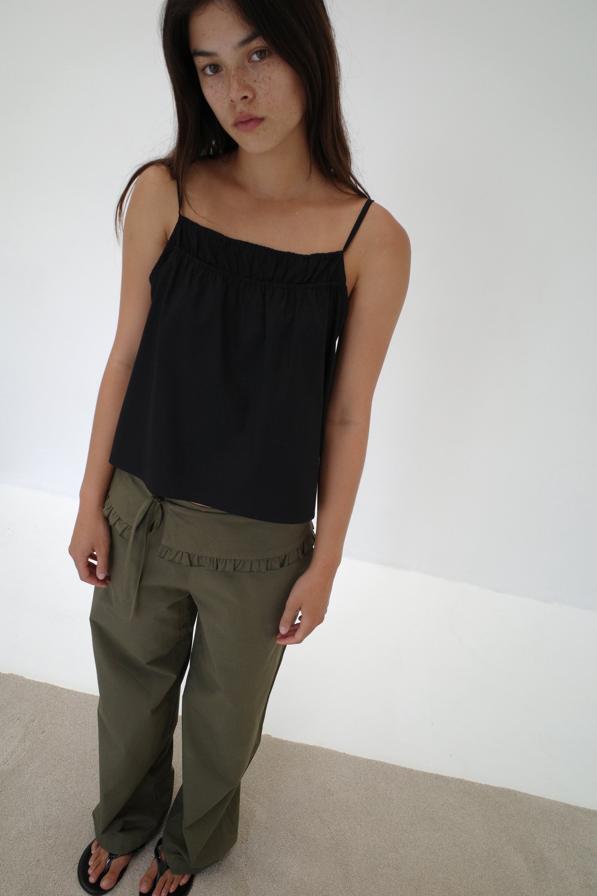 A woman with long brown hair wears a loose black spaghetti strap top, the Flounce Belt Pant in khaki, and black sandals, standing indoors against a plain white wall and beige floor, gazing softly at the camera.