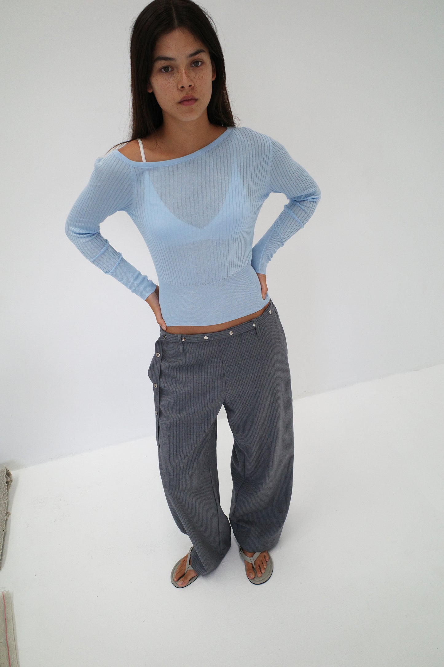A woman with long dark hair stands against a white background, wearing the Deiji Studios Shoulder Knit Top in blue and loose gray pinstriped pants with buttons, paired with sandals. She faces the camera.