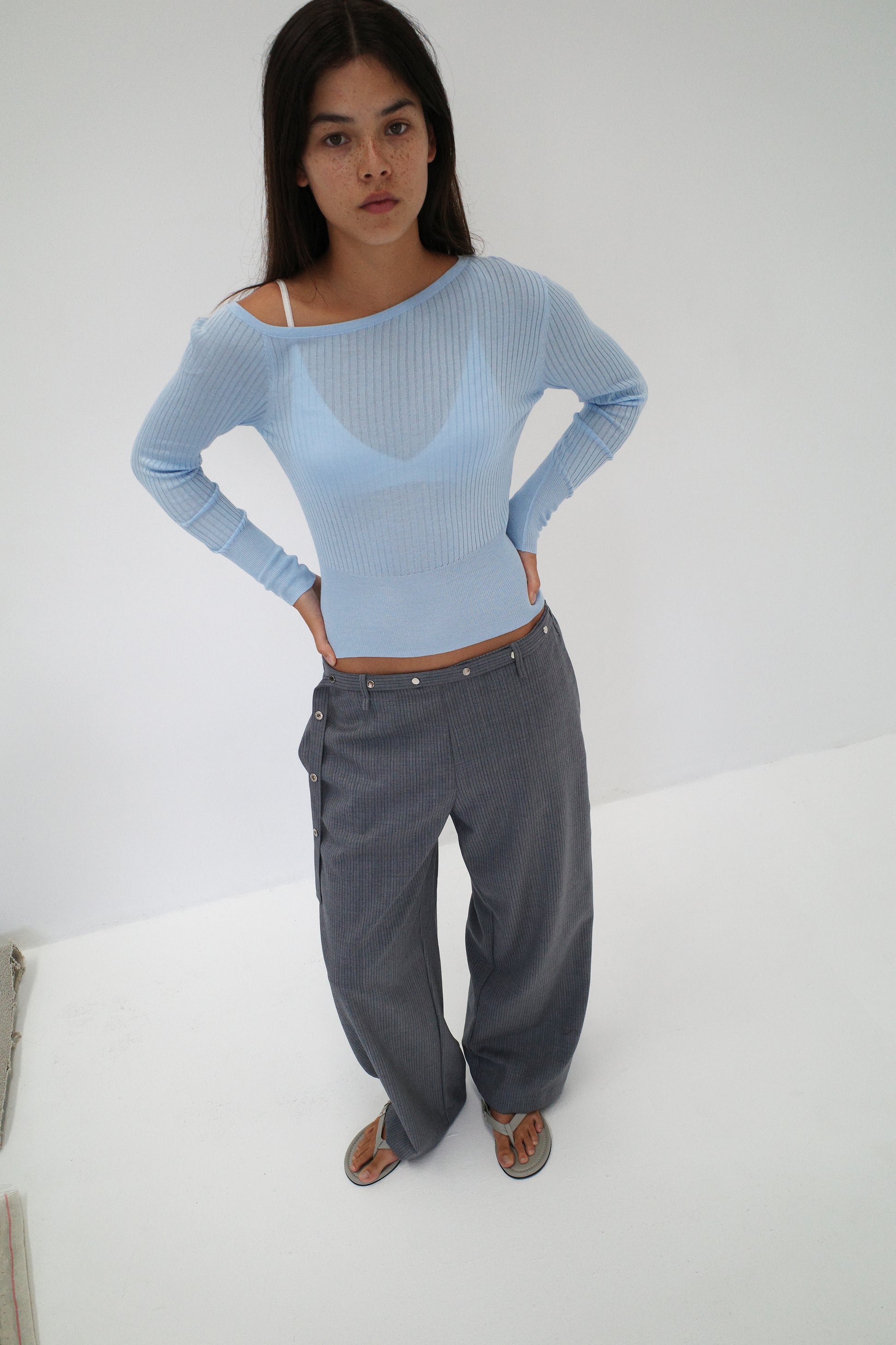 A woman with long dark hair stands against a white background, wearing the Deiji Studios Shoulder Knit Top in blue and loose gray pinstriped pants with buttons, paired with sandals. She faces the camera.
