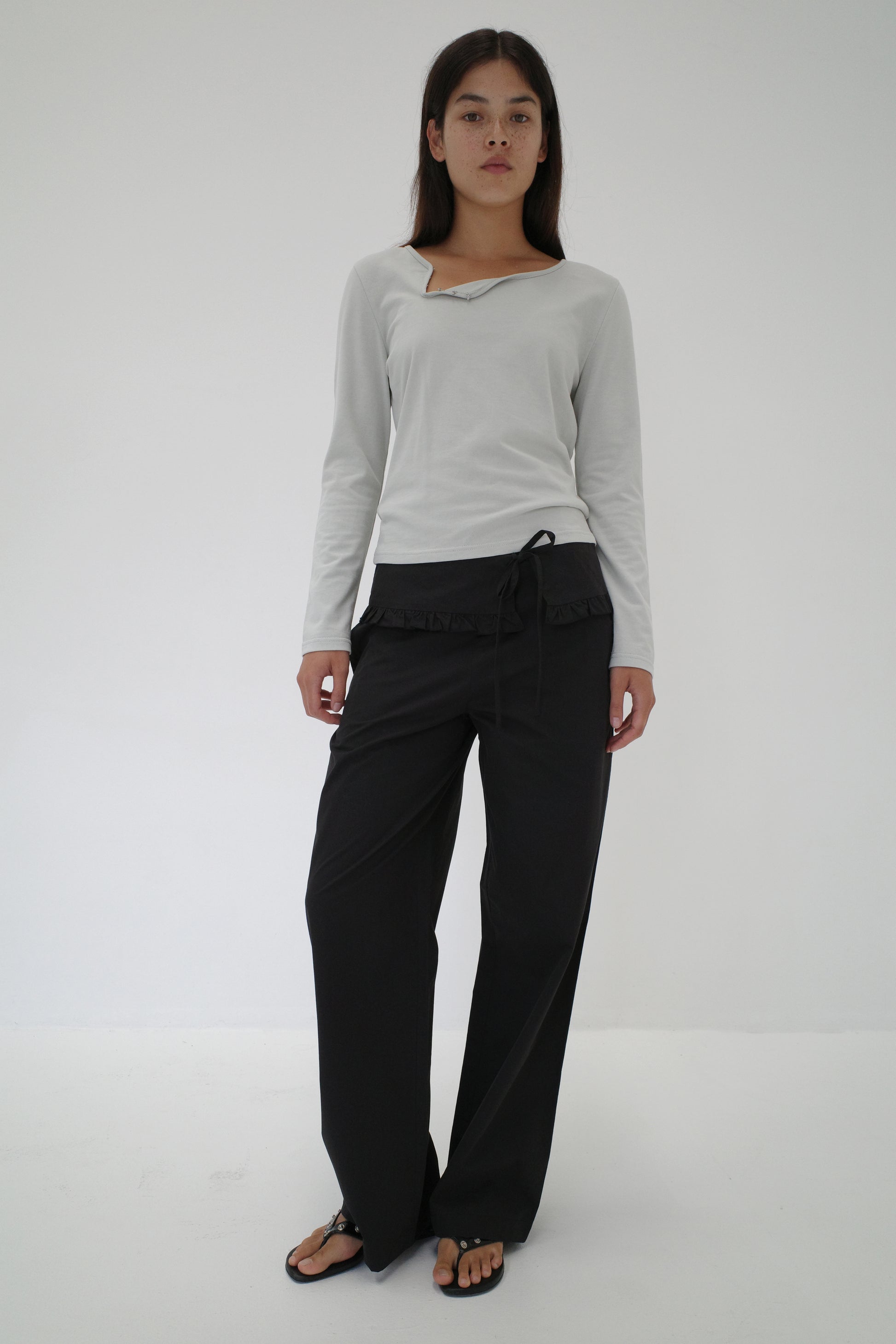 A woman stands against a plain white background wearing a light gray long-sleeve top, Flounce Belt Pant - Black with a tie at the waist, and black sandals. She looks straight ahead with a neutral expression.