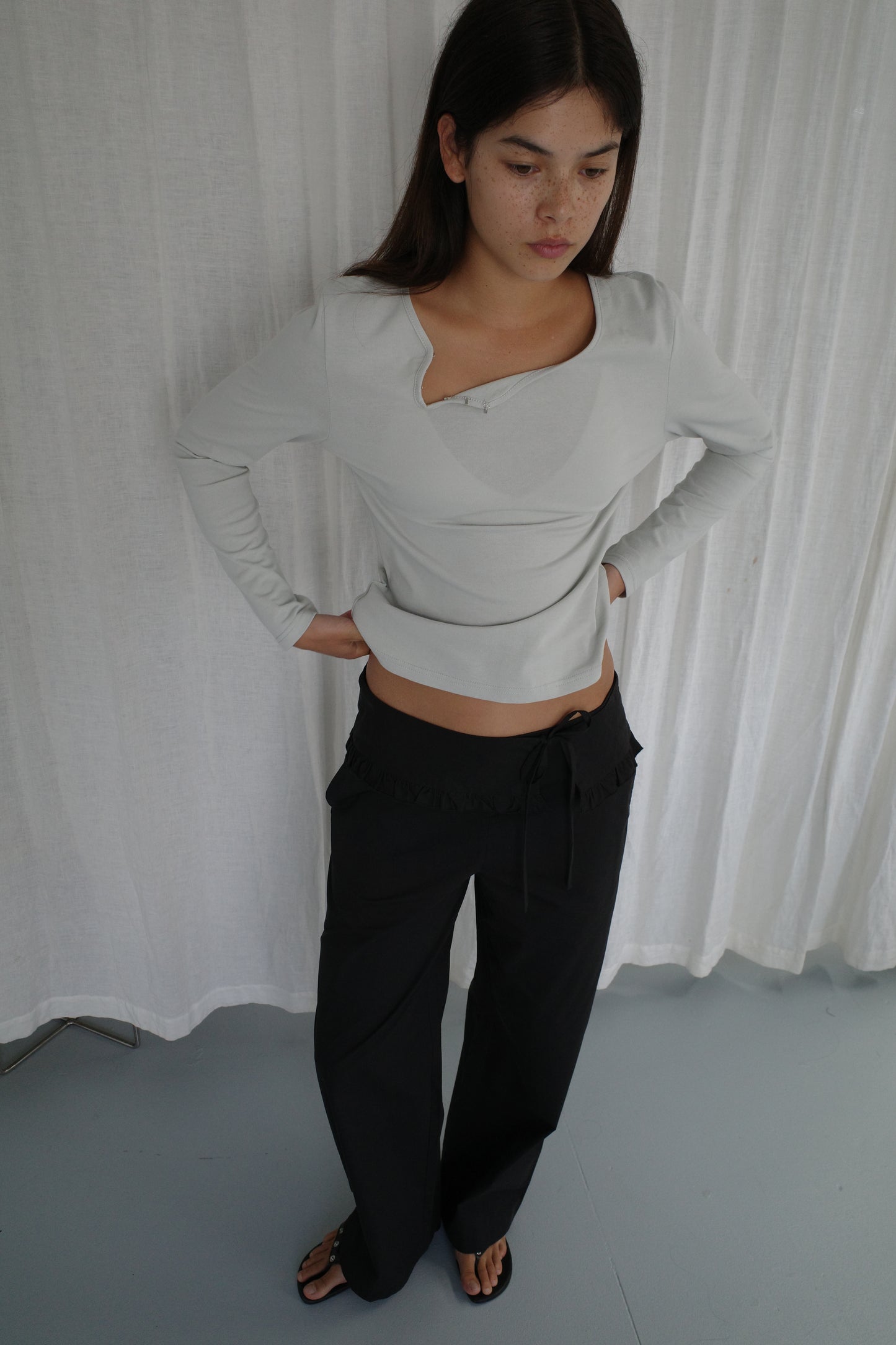 A young woman with long dark hair stands before a white curtain in a light gray asymmetric top and Flounce Belt Pant - Black, hands on hips as she looks down, highlighting the pant’s relaxed yet refined silhouette.