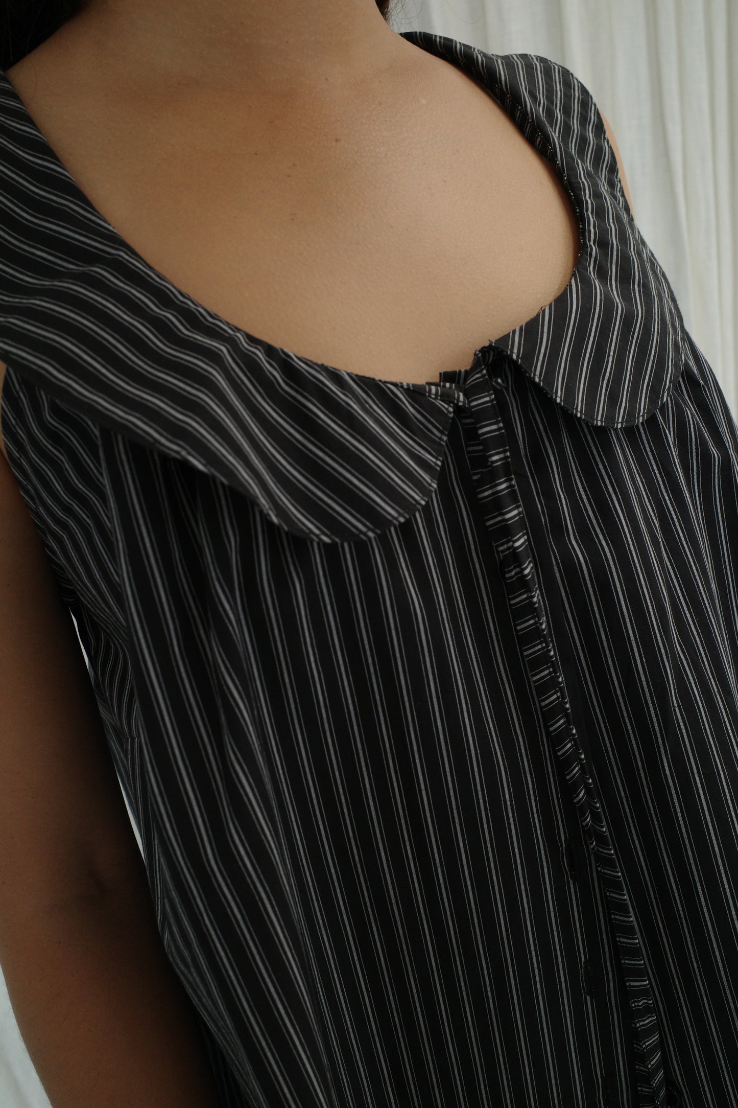 Close-up of a person wearing the Round Collar Top - Mono Stripe by Deiji Studios, a sleeveless top with white vertical pinstripes, large collar, and front buttons. The softly lit background features white curtains.