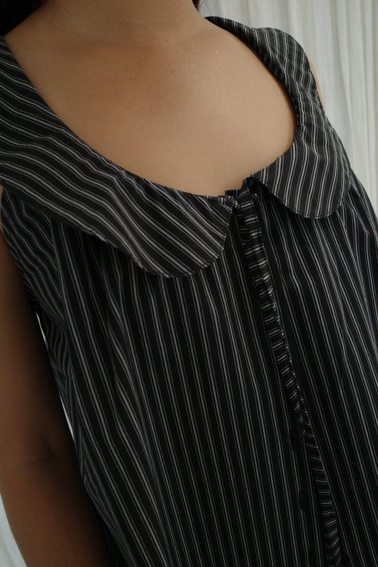 Close-up of a person wearing the Round Collar Top - Mono Stripe by Deiji Studios, a sleeveless top with white vertical pinstripes, large collar, and front buttons. The softly lit background features white curtains.