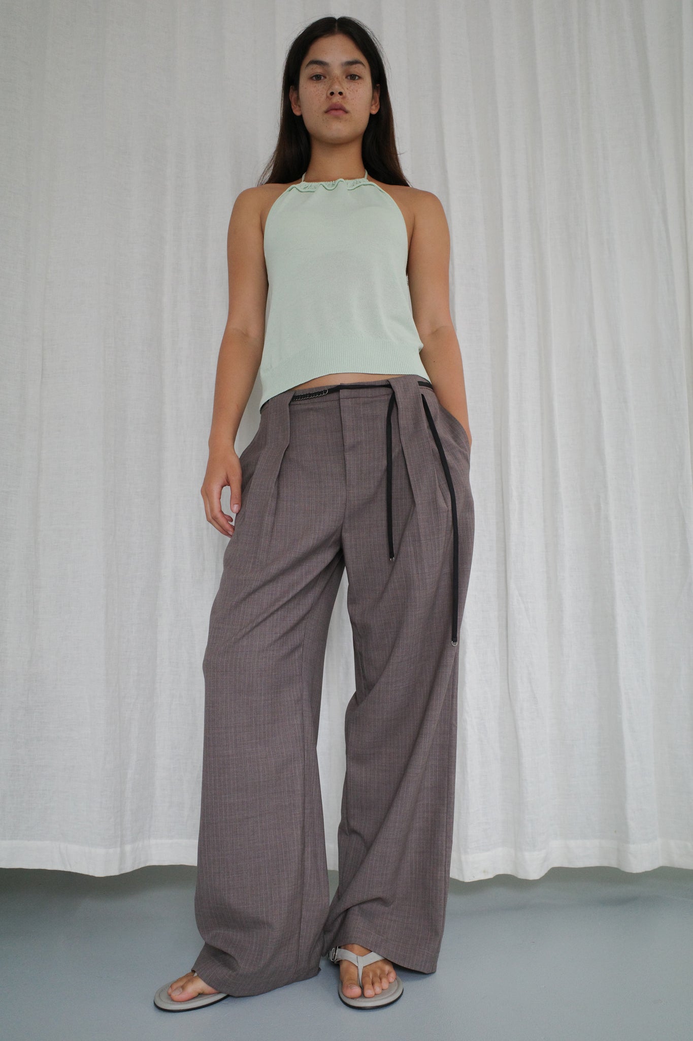 A woman stands in front of white curtains wearing a light green halter top, Belt Loop Pant - Grey Pink Stripe, and gray sandals. She looks confidently at the camera.