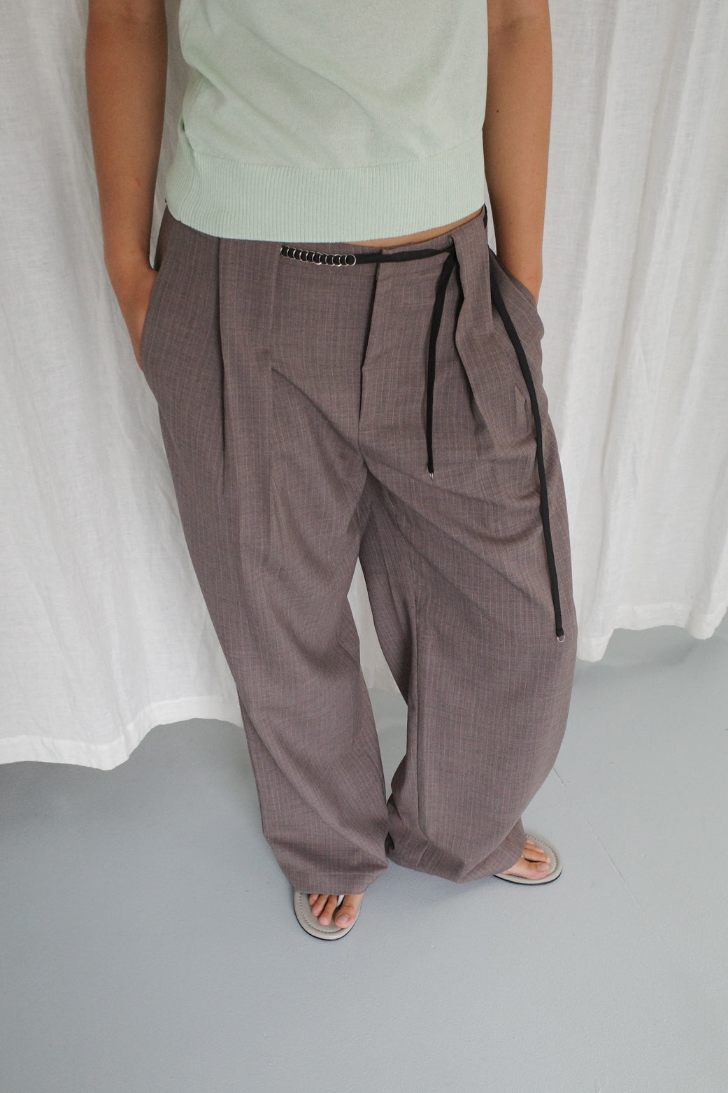 A person wears Belt Loop Pant - Grey Pink Stripe, styled with a light green top and sandals, standing against a white fabric backdrop with hands in their pockets.