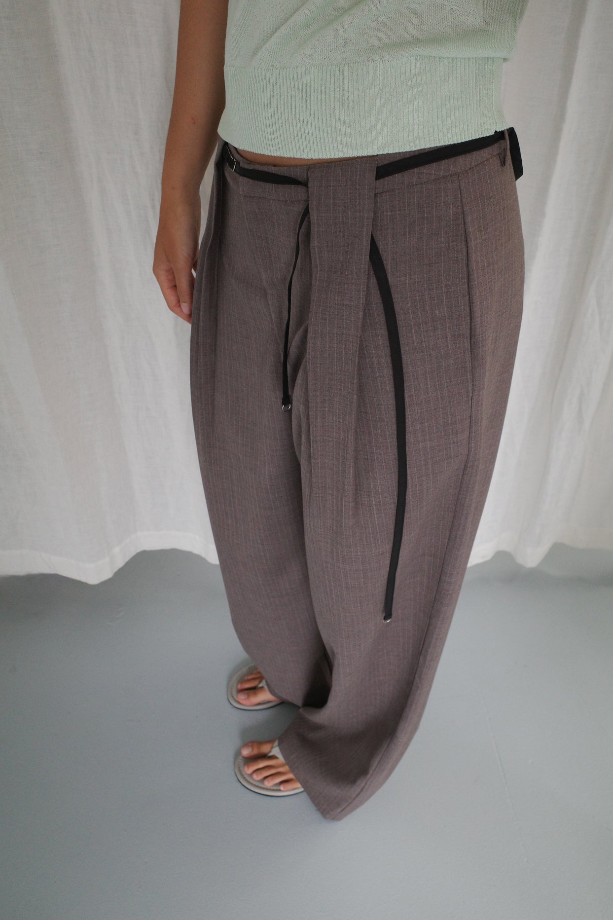 A person stands indoors wearing Belt Loop Pant - Grey Pink Stripe, paired with a light green top and gray open-toe sandals, against a backdrop of a white curtain and light gray floor.