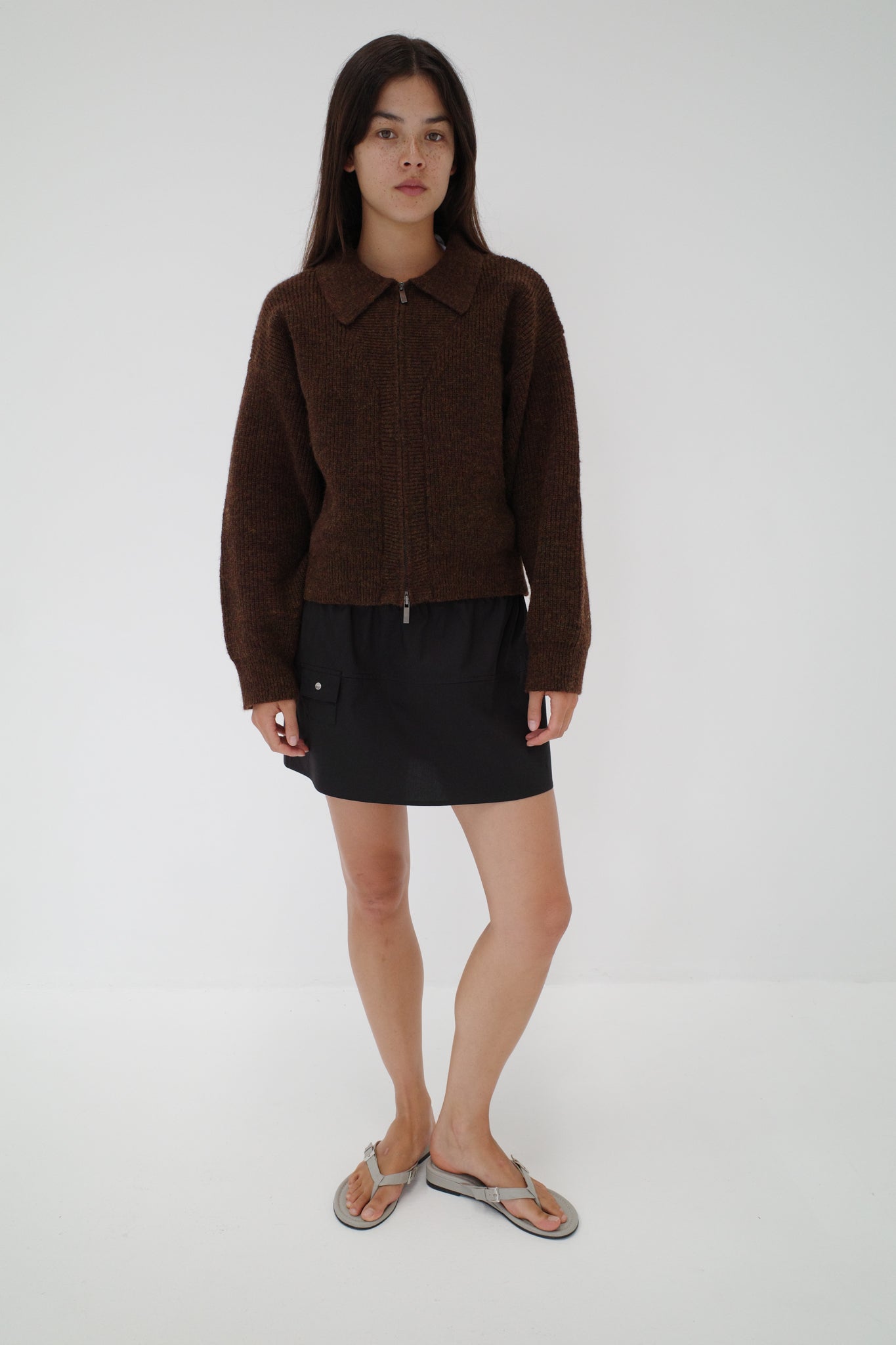 A woman with long dark hair and a neutral expression stands against a white background wearing Deiji Studios’ Double Layer Cardigan in brown, paired with a black mini skirt and silver flip-flops.