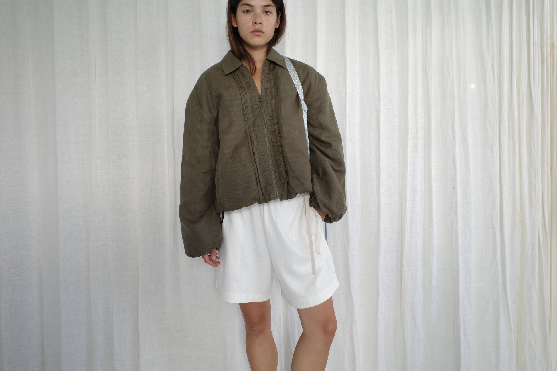 A person stands against a white curtain, wearing the Pintuck Jacket - Khaki Stripe from Deiji Studios over a white outfit with shorts, looking directly at the camera with a neutral expression.