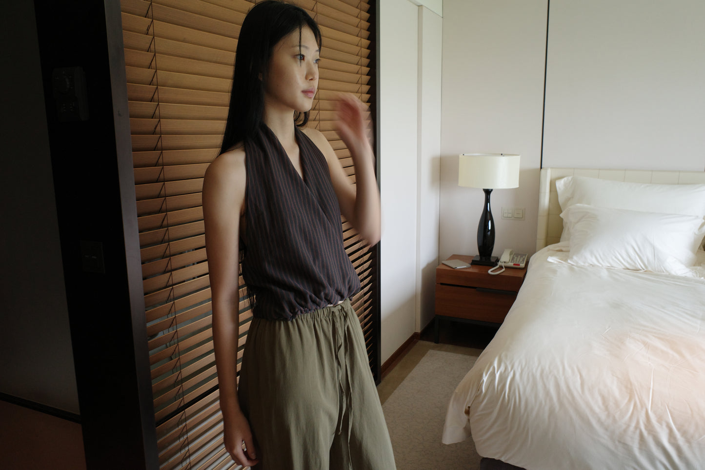 A woman with long dark hair stands beside wooden blinds in a bedroom wearing the Wrap Halter Top - Brown Grey Stripe by Deiji Studios, a sheer brown and grey striped halter top, with khaki trousers.