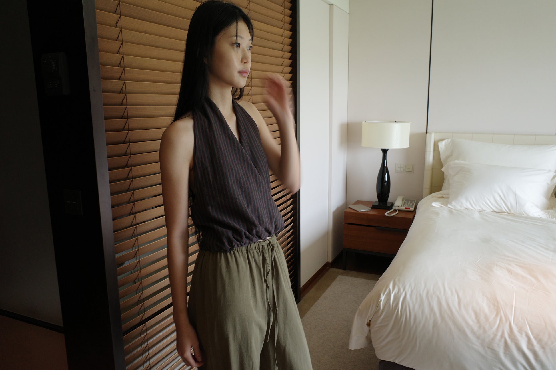 A woman with long dark hair stands beside wooden blinds in a bedroom wearing the Wrap Halter Top - Brown Grey Stripe by Deiji Studios, a sheer brown and grey striped halter top, with khaki trousers.
