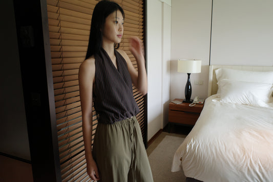 A woman with long dark hair stands beside wooden blinds in a bedroom wearing the Wrap Halter Top - Brown Grey Stripe by Deiji Studios, a sheer brown and grey striped halter top, with khaki trousers.