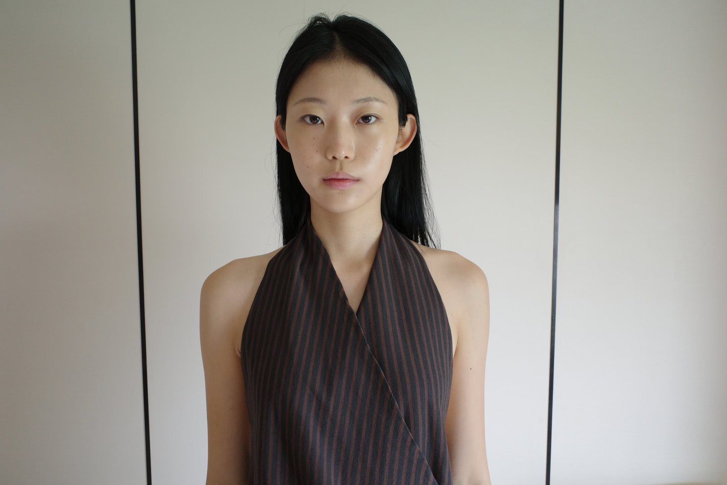 A cropped front view of a woman wearing the Wrap Halter Top - Brown Grey Stripe by Deiji Studios, a sheer brown and grey striped halter top with a wrap surplice front, against a pale wall.