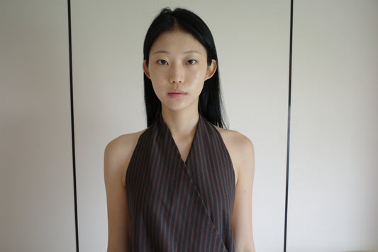 A cropped front view of a woman wearing the Wrap Halter Top - Brown Grey Stripe by Deiji Studios, a sheer brown and grey striped halter top with a wrap surplice front, against a pale wall.
