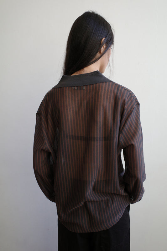 A woman with long dark hair stands with her back to the camera against a pale wall wearing the Knit Shirt - Brown Grey Stripe by Deiji Studios, a sheer brown and charcoal striped knit shirt with a dark collar.