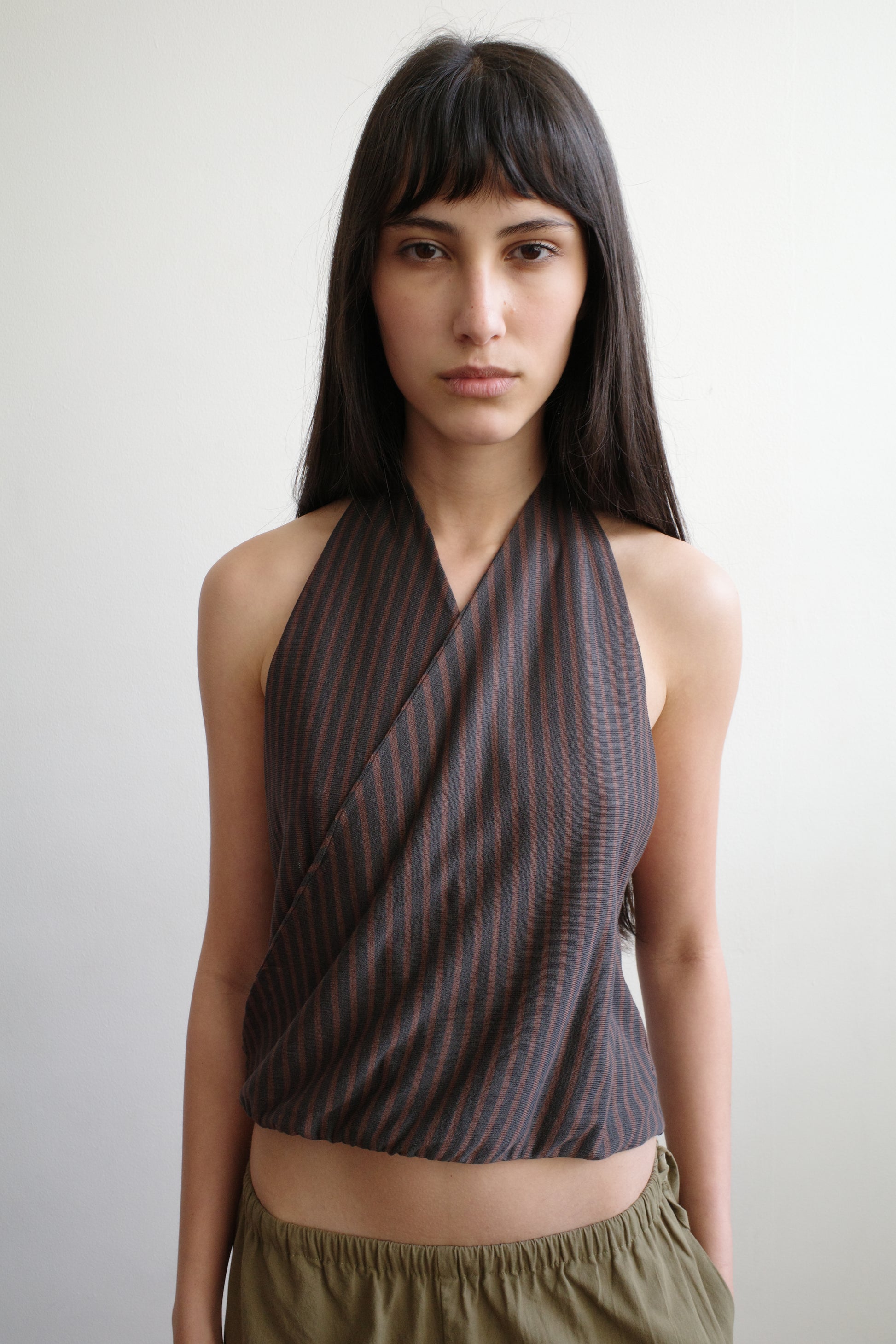 A woman with long dark hair and bangs wears the Wrap Halter Top in Brown Grey Stripe from Deiji Studios, paired with olive green pants, standing against a plain white background.