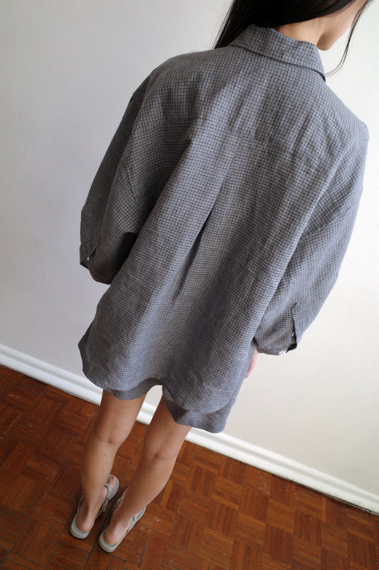 A woman with long dark hair looks to the side, standing in a room and wearing the 03 Set - Indigo Check by Deiji Studios, an oversized boxy check linen shirt with matching boxer-style shorts.