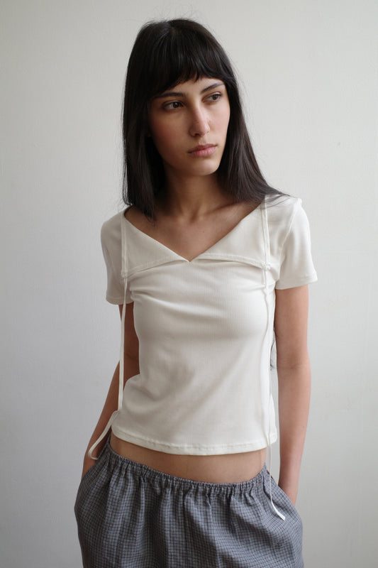 A woman with dark hair and a fringe stands against a pale wall wearing the Tie Back Tee - White by Deiji Studios, a fitted cream tee with a sweetheart neckline and back ties, with grey check trousers.