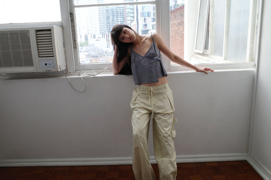 A woman with dark hair and a fringe leans against a window sill wearing the Wrapping Top - Indigo Check by Deiji Studios, an indigo micro-check linen cami, with cream trousers.