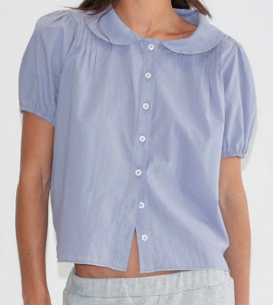 A person wearing the Petal Top, a light blue short-sleeve button-up blouse with a Peter Pan collar, paired with grey pants and shown from shoulders to waist.