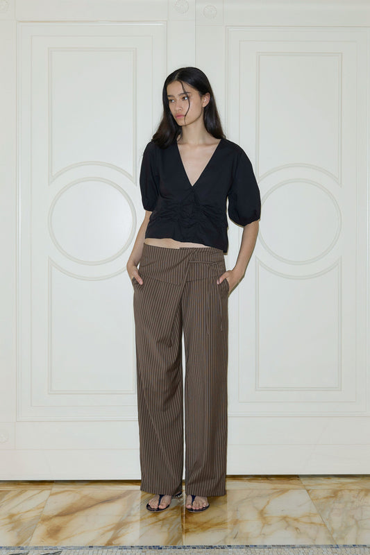 A woman with long dark hair stands indoors in a black organic cotton Draw Top with puff sleeves and a V-neck, paired with high-waisted brown pinstriped wide-leg trousers and black sandals. She poses with one hand in her pocket.