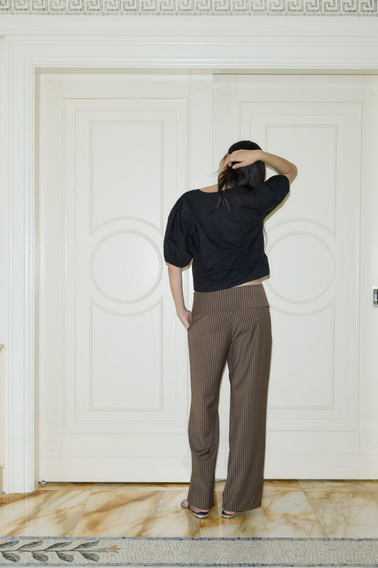 A person stands indoors facing a large double door, back to the camera, wearing the Draw Top - Black, brown pinstripe pants, and sandals, with one hand on their head and the other by their side.