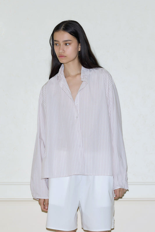 A person with long dark hair wears the Deiji Studios day shirt—a loose, light pink and white striped button-up—paired with white shorts, standing against a plain, light-colored wall.