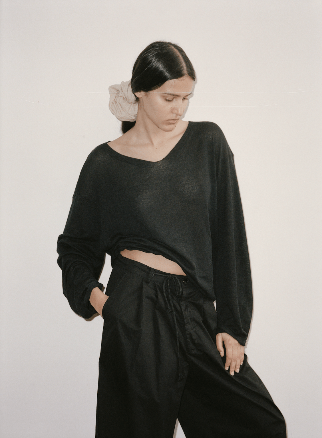 Female model wears the Deiji Studios Loose Long Sleeve Knitted Top in black, styled with the Cotton Pant in black and Scrunchie in Sand Stripe worn in hair. Knit top shows light, open weave and subtle woven neckline detail.