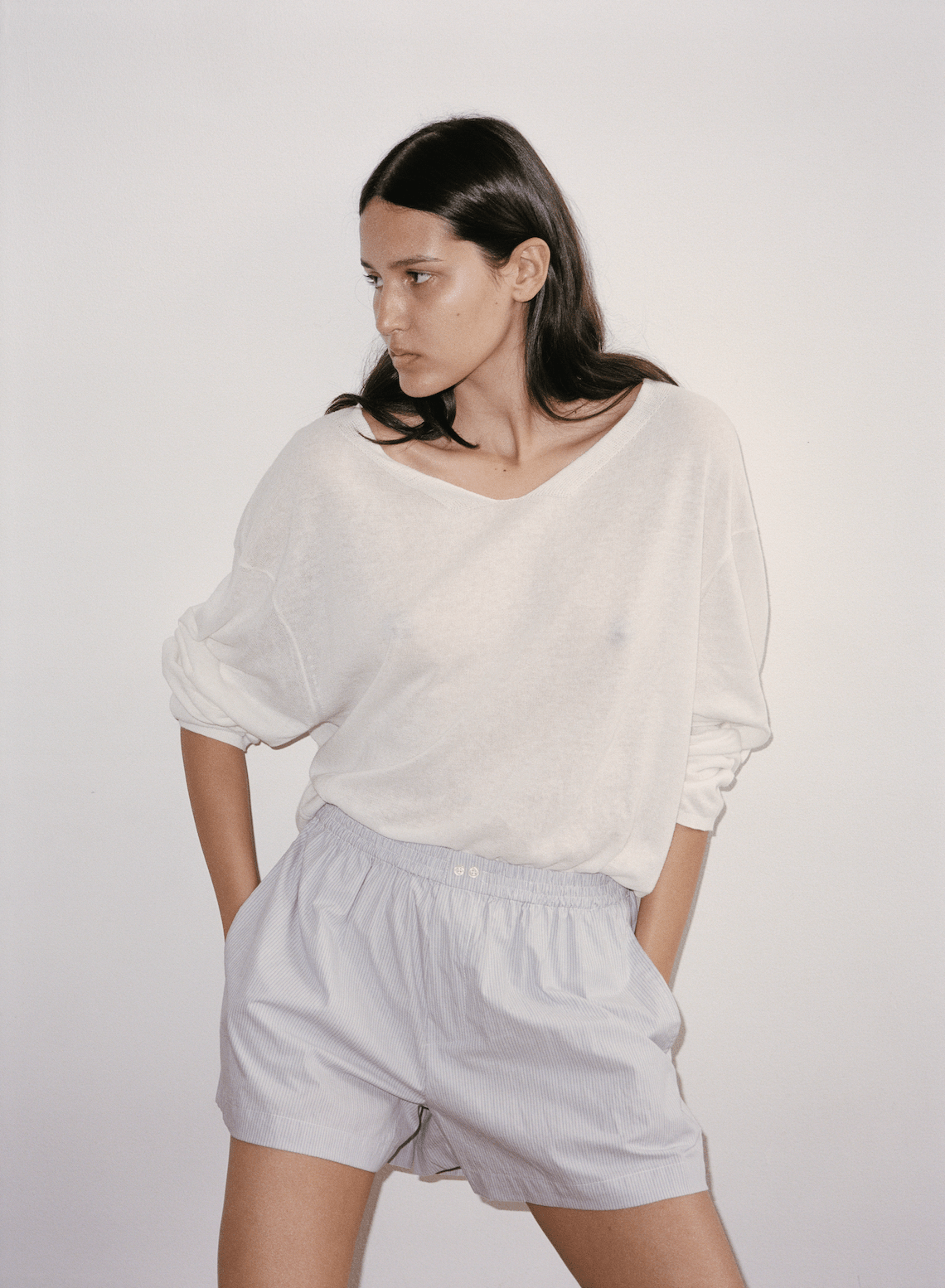 Film image of female model leaning against wall wearing the Loose Long Sleeve Knitted Top by Deiji Studios in white, paired with the Boxer in Dream Stripe.