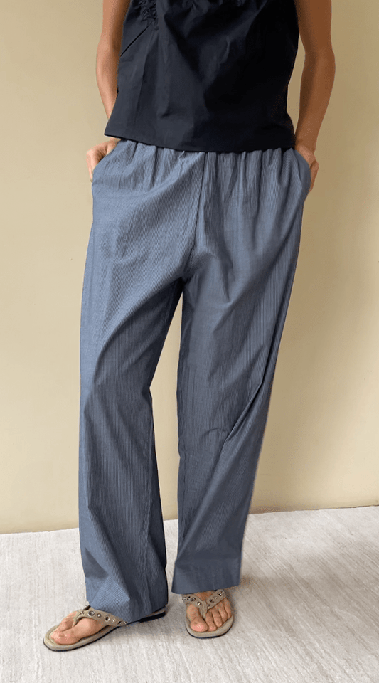 Wearing the Ease Trouser - Blue Pinstripe, made from organic cotton with a relaxed fit and elastic waist, paired with a sleeveless black top and beige sandals, the person stands with hands in pockets against a plain background.