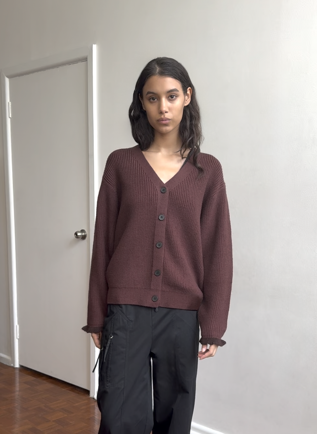 A woman with long dark hair stands indoors, wearing the Flounce Cardigan - Burgundy and black pants. She faces the camera in a minimalist room with white walls and a closed door.