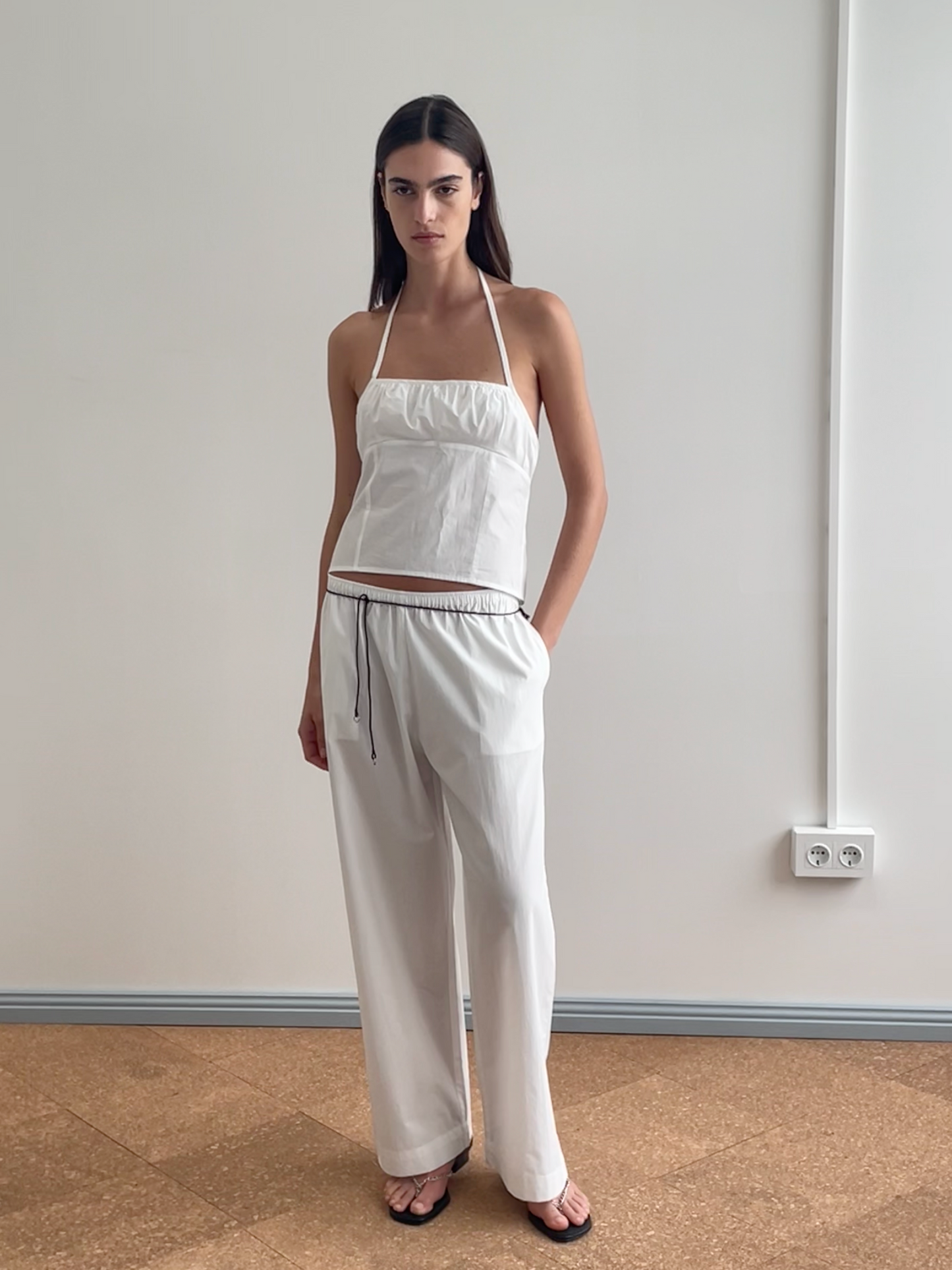 A woman with straight dark hair and a neutral expression stands indoors against a plain wall, wearing the Strap Top - White (an organic cotton halter top), loose white drawstring pants, and black sandals.