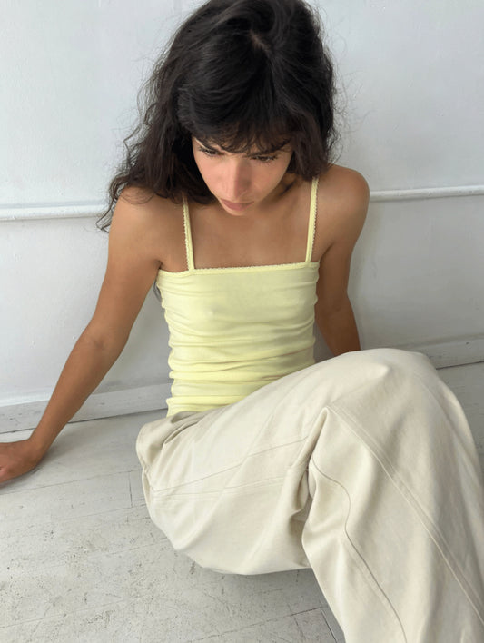 A woman sits on a white studio floor, wearing the Scallop Tank - Lemon by Deiji Studios, a fitted ribbed tank with fine shoulder straps and scallop trim along the straight neckline.