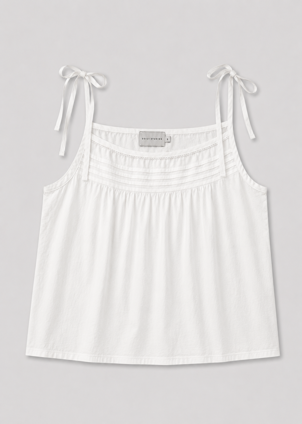 The Tied Cami - White by Deiji Studios features shoulder tie straps and subtle chest pleating, shown on a plain light gray background.