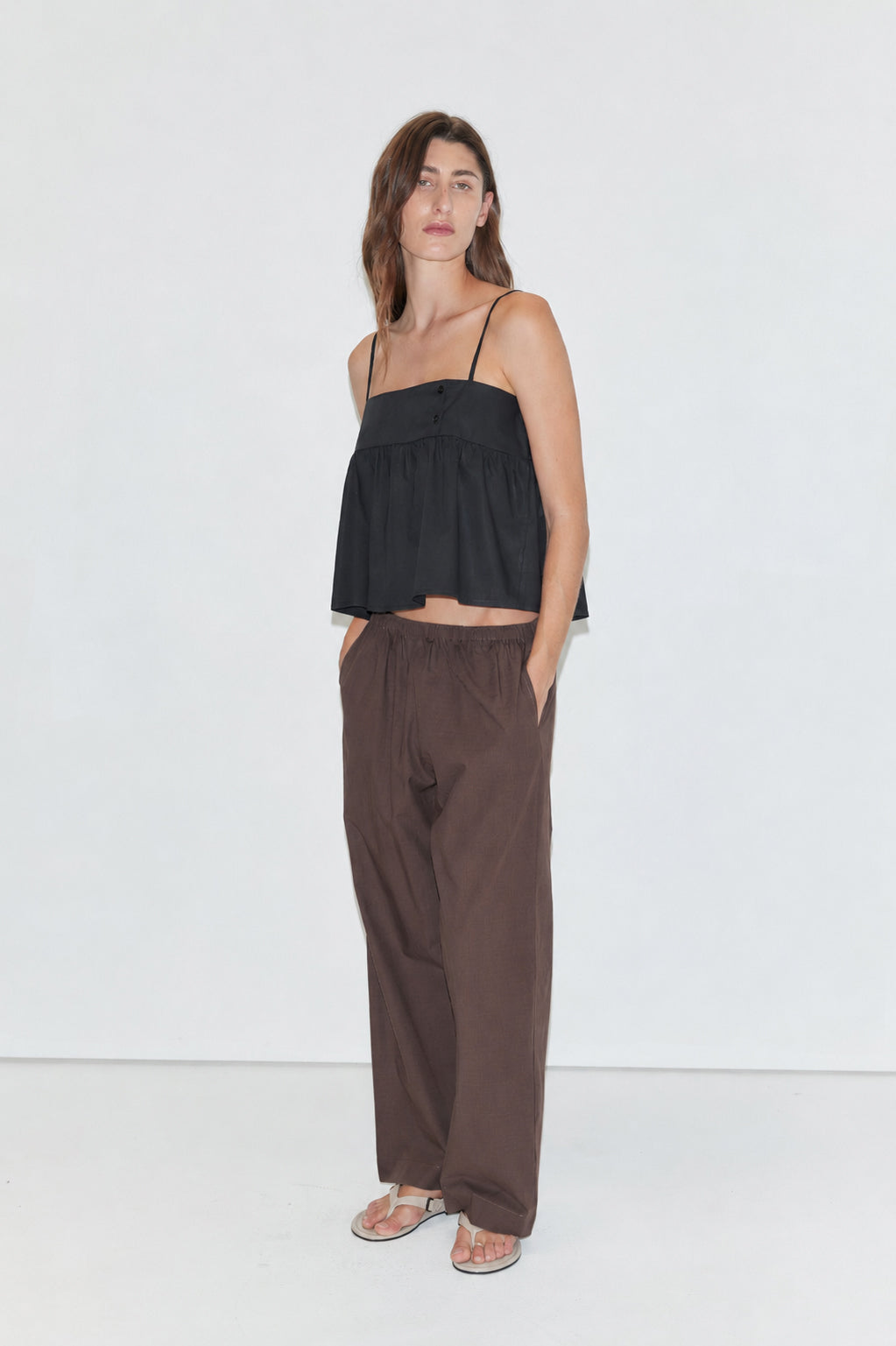A woman stands against a plain white background wearing a black spaghetti-strap top, Ease Trouser - Toast by Deiji Studios, and sandals, with her hands in her pockets and a neutral expression.