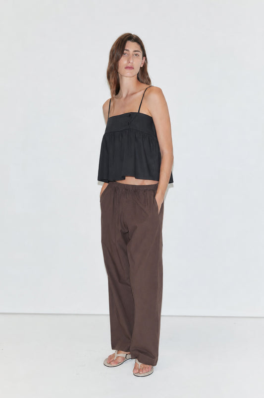 A woman stands against a plain white background wearing a black spaghetti-strap top, Ease Trouser - Toast by Deiji Studios, and sandals, with her hands in her pockets and a neutral expression.