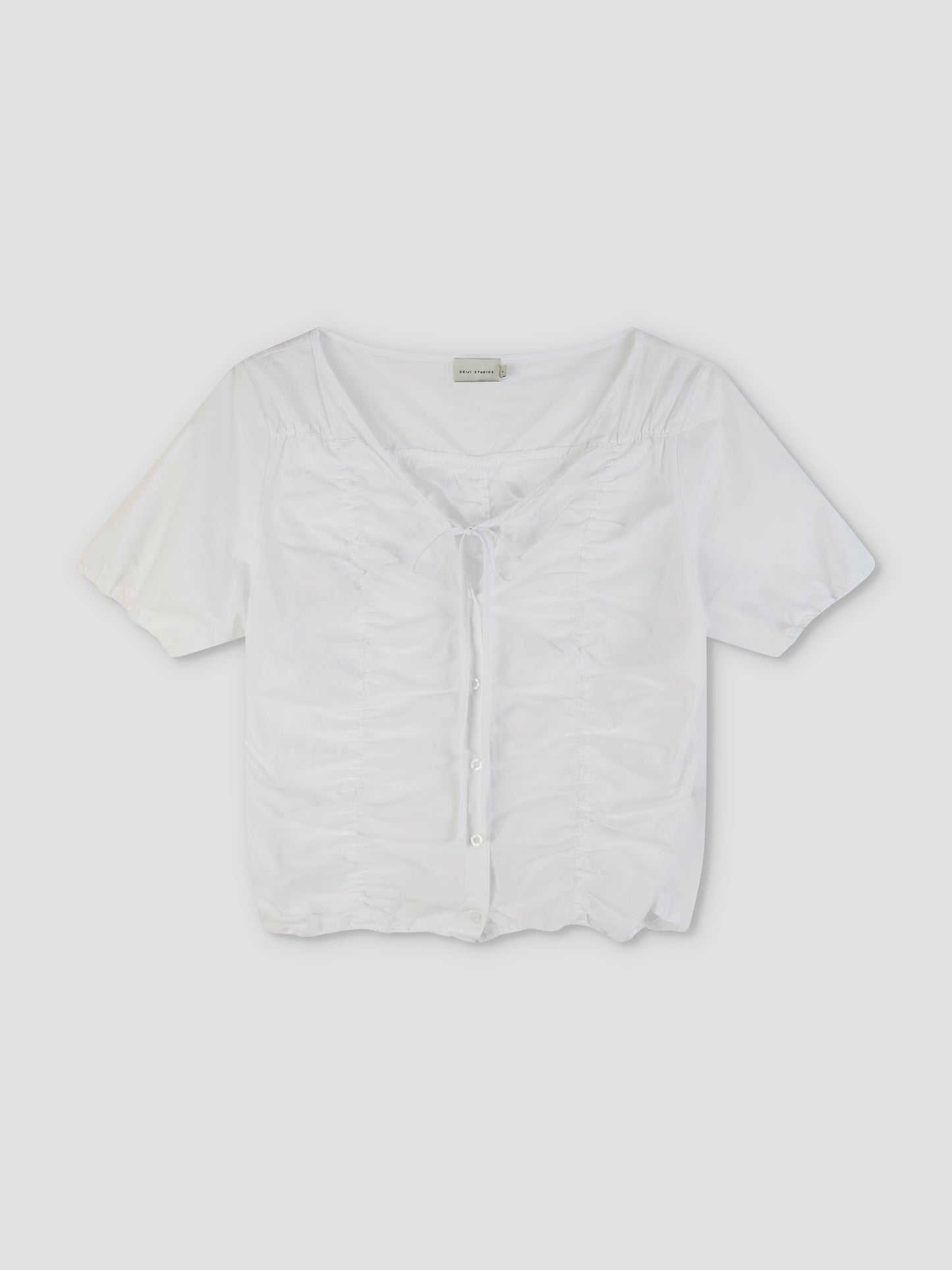 The Crinkle Top - White is a short-sleeved, white button-up top featuring a ruched front, tie detail at the neckline, and a slightly cropped hem, displayed against a plain light background.