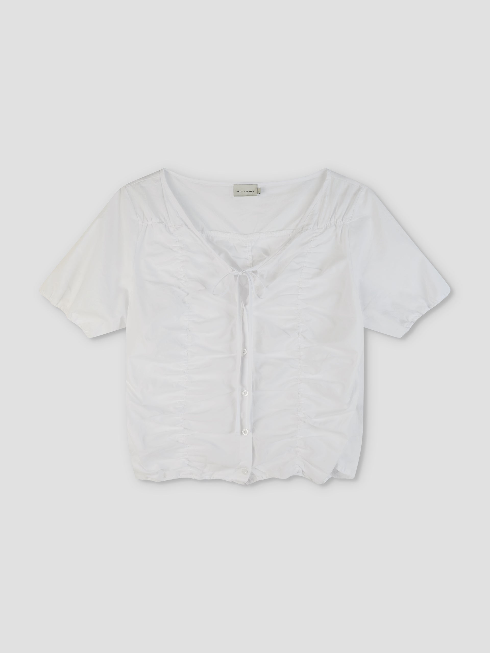 The Crinkle Top - White is a short-sleeved, white button-up top featuring a ruched front, tie detail at the neckline, and a slightly cropped hem, displayed against a plain light background.