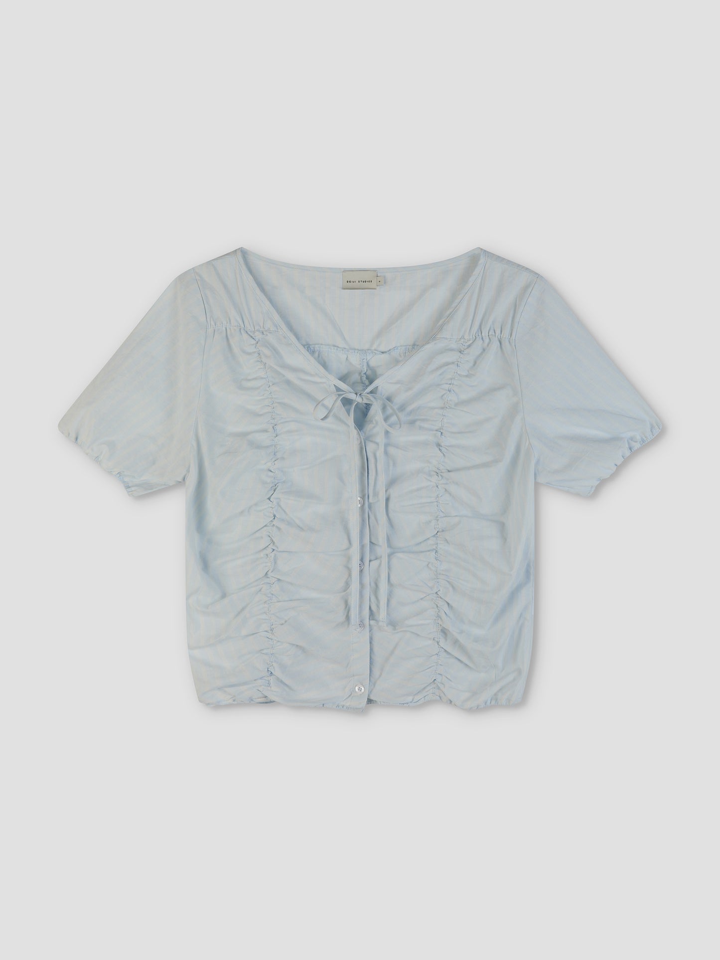 The Crinkle Top - Powder Blue Stripe features a V-neckline, ruched front with center tie, button-down hem, and lightweight sheer fabric in a softly textured blue stripe, reflecting Deiji Studios' relaxed style. Shown laid flat on a plain background.