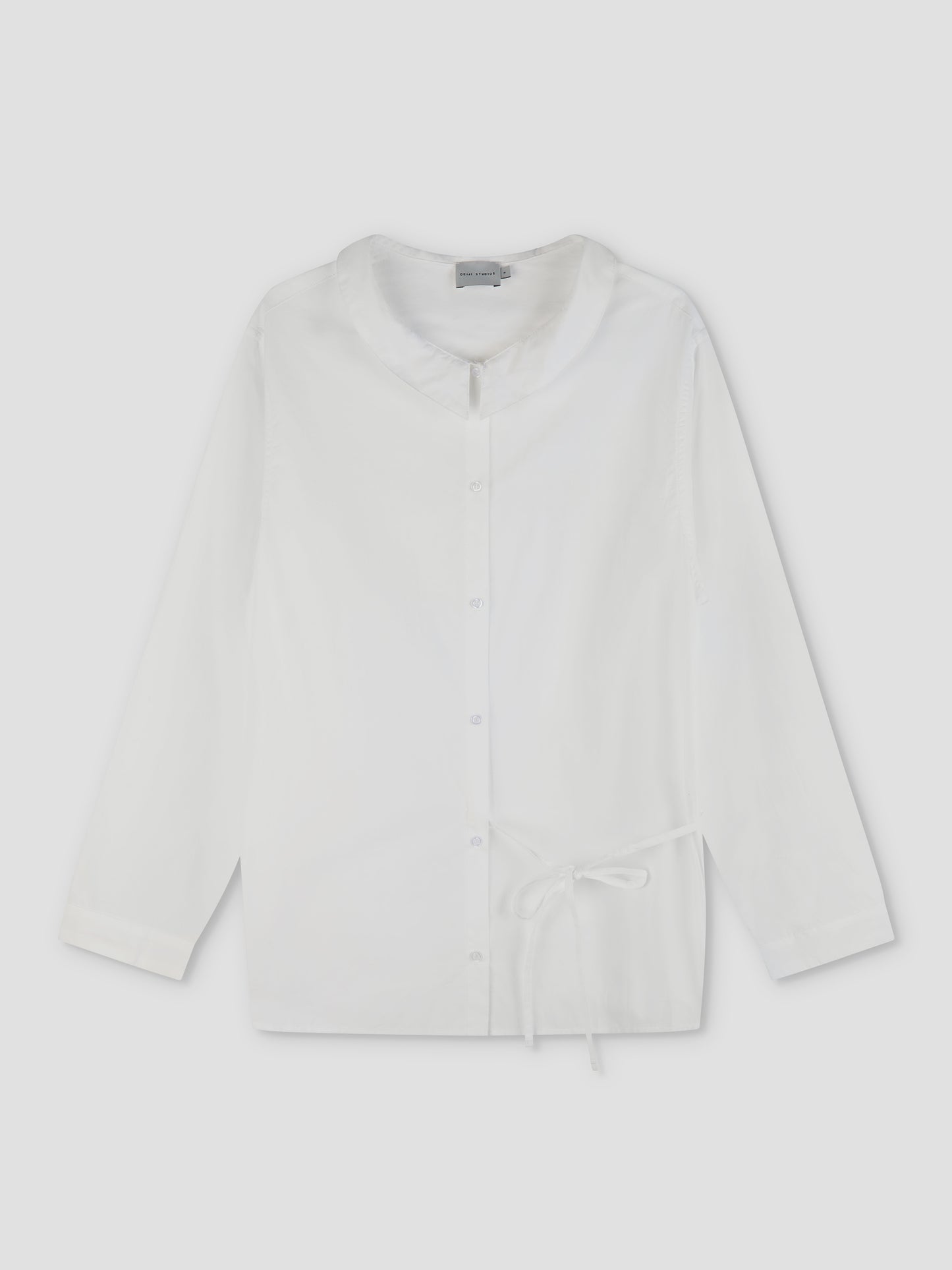 The Left Shirt - White is a lightweight, long-sleeve button-up with a rounded collar, relaxed fit, and an adjustable bow side tie at the waist, shown flat on a light background.