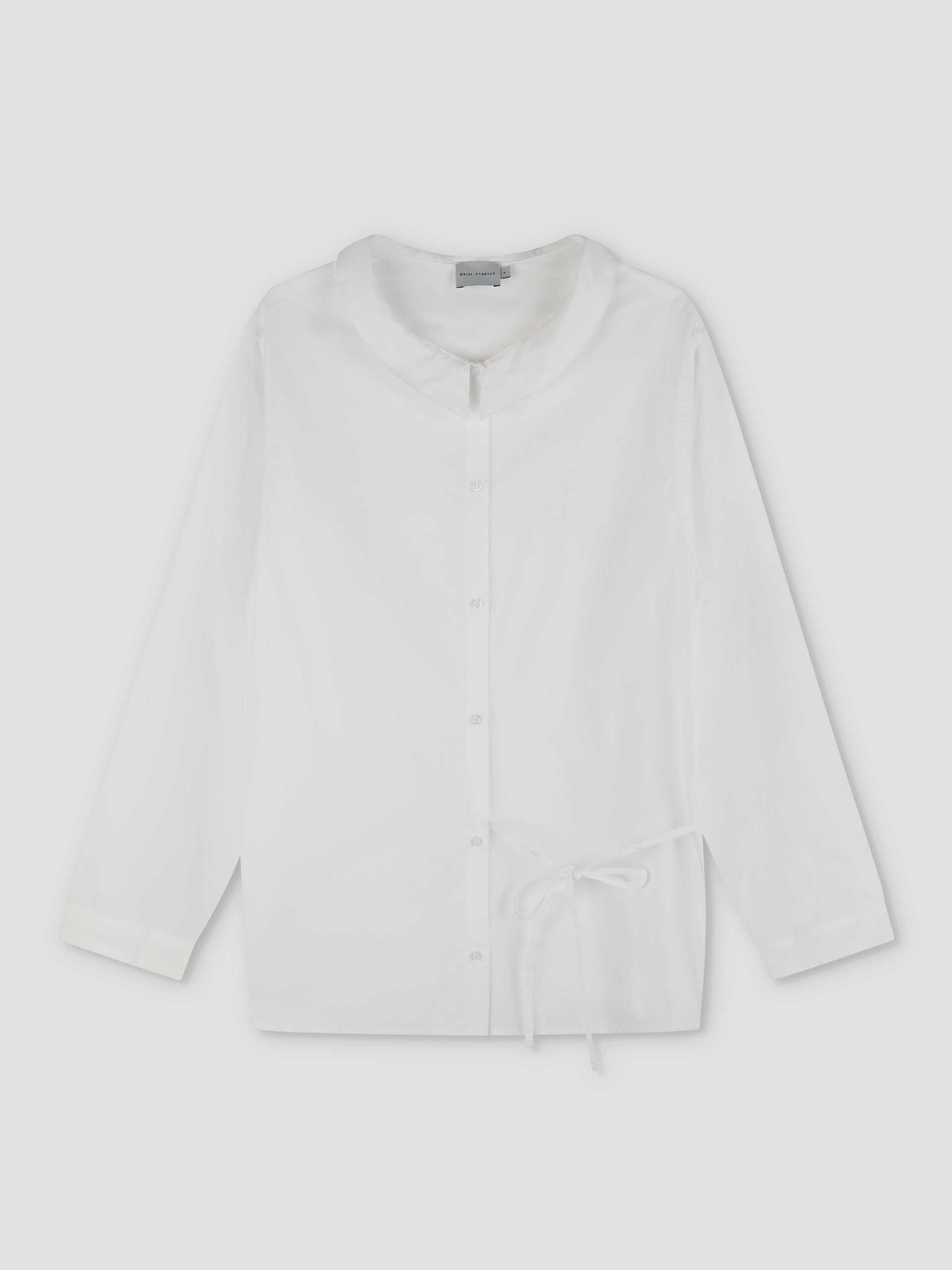 The Left Shirt - White is a lightweight, long-sleeve button-up with a rounded collar, relaxed fit, and an adjustable bow side tie at the waist, shown flat on a light background.