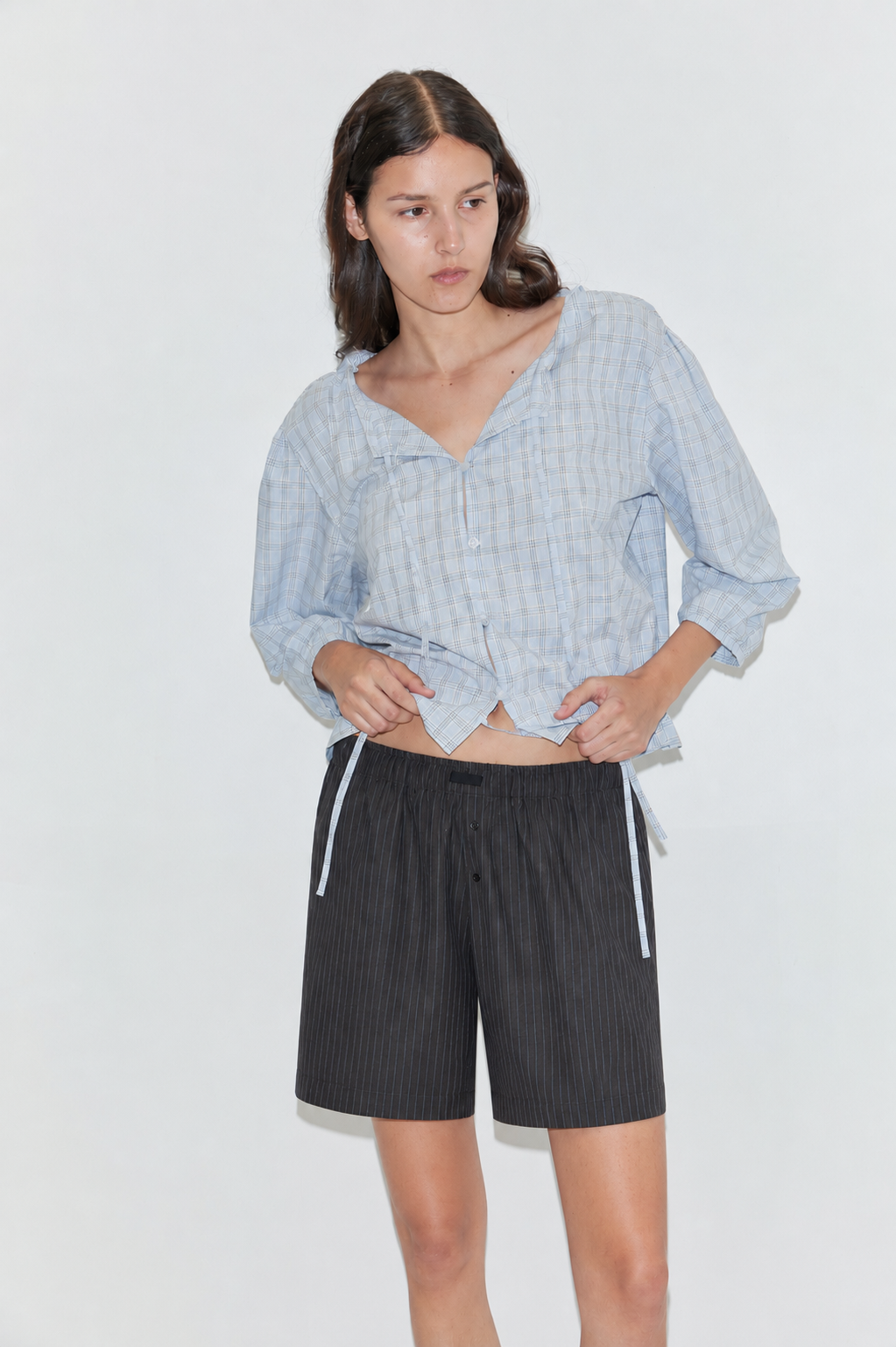 A woman stands against a plain light background, wearing the Corded Top - Powder Check from Deiji Studios and black pinstripe shorts. She looks slightly to the side with one hand resting on her hip.