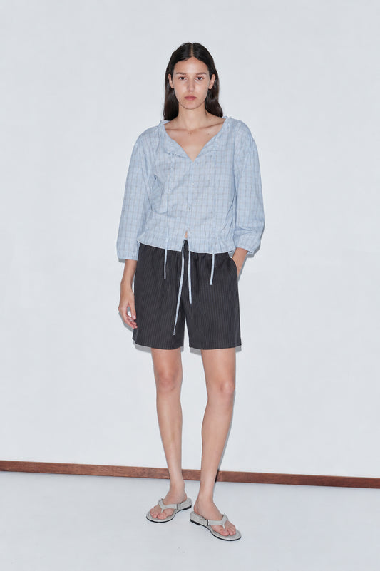 A woman with long dark hair and a neutral expression wears a light blue v-neck blouse, Wool Boxer Short - Charcoal Stripe by Deiji Studios, and flip-flops against a plain background.