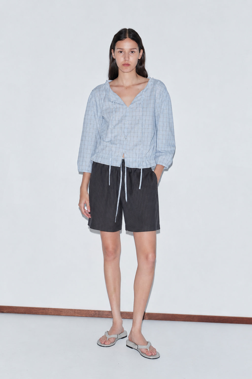 A woman with long dark hair and a neutral expression stands against a plain white background wearing the Corded Top - Powder Check by Deiji Studios, dark pinstriped shorts, and flip-flops.