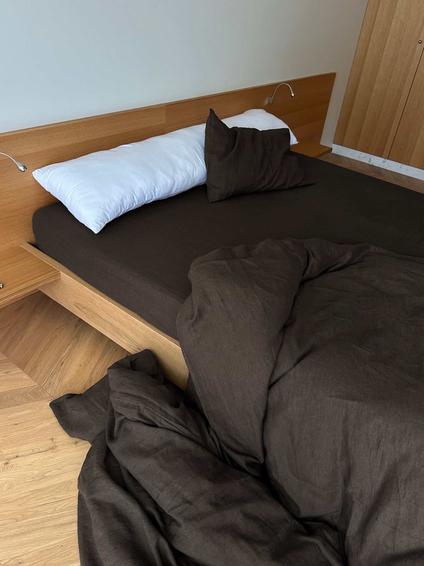 A modern wooden bed with a Duvet cover - Bark, dark brown blanket partially on the floor, white cylindrical pillow, and breathable sheets. Wooden floors and light walls complete the cozy unmade look.