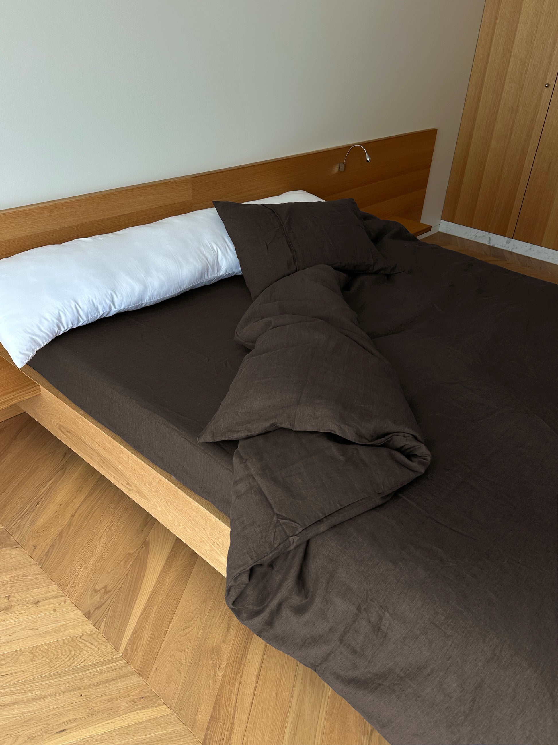 A modern wooden bed with a white pillow and dark brown hypo-allergenic Duvet cover - Bark rests on a light wood floor. The comforter is slightly messy, with a built-in headboard and wardrobe in the background.