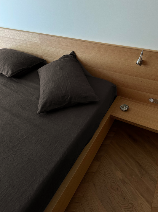 A neatly made bed features dark brown sheets and two pillows in the Pillow slip set - Bark, made from organic linen. The wooden headboard has a silver reading light and a bedside shelf with a stone.