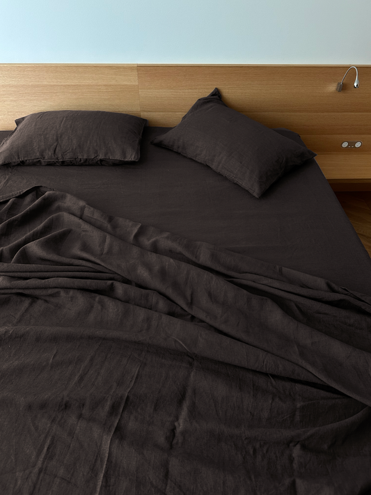 A bed with hypo-allergenic Sheets - Bark in dark brown and two pillows sits against a wooden headboard, which has a wall-mounted reading light and power outlets. The sheets are slightly rumpled.